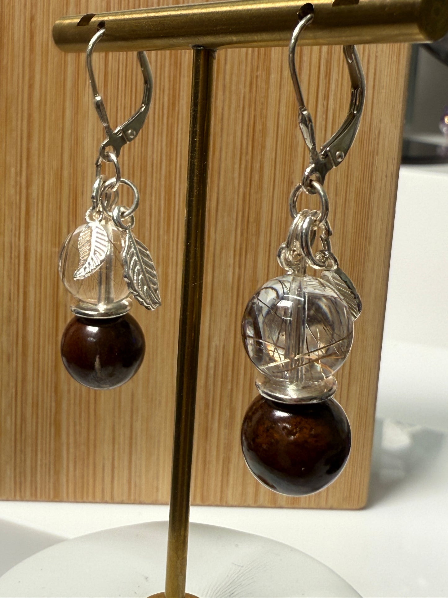 Close-up of a pair of crystal earrings featuring Rutile-in-Quartz and Brown Opal stones, accented with silver leaf charms. Displayed against a soft neutral background.