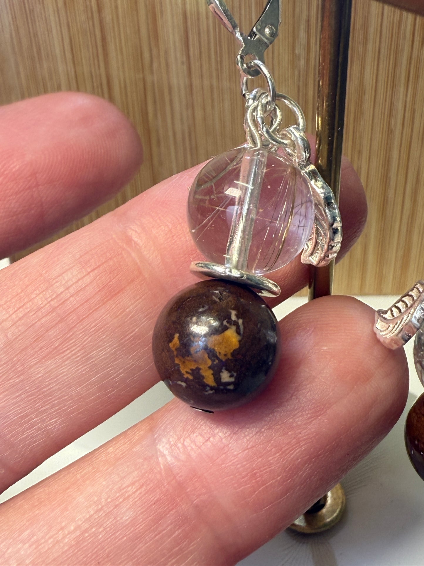 Close-up of a pair of crystal earrings featuring Rutile-in-Quartz and Brown Opal stones, accented with silver leaf charms. Displayed against a soft neutral background.