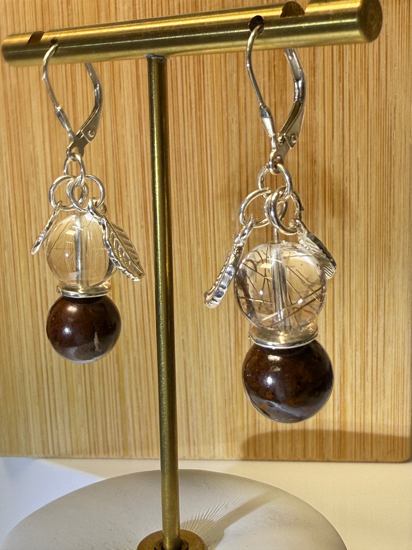 Close-up of a pair of crystal earrings featuring Rutile-in-Quartz and Brown Opal stones, accented with silver leaf charms. Displayed against a soft neutral background.